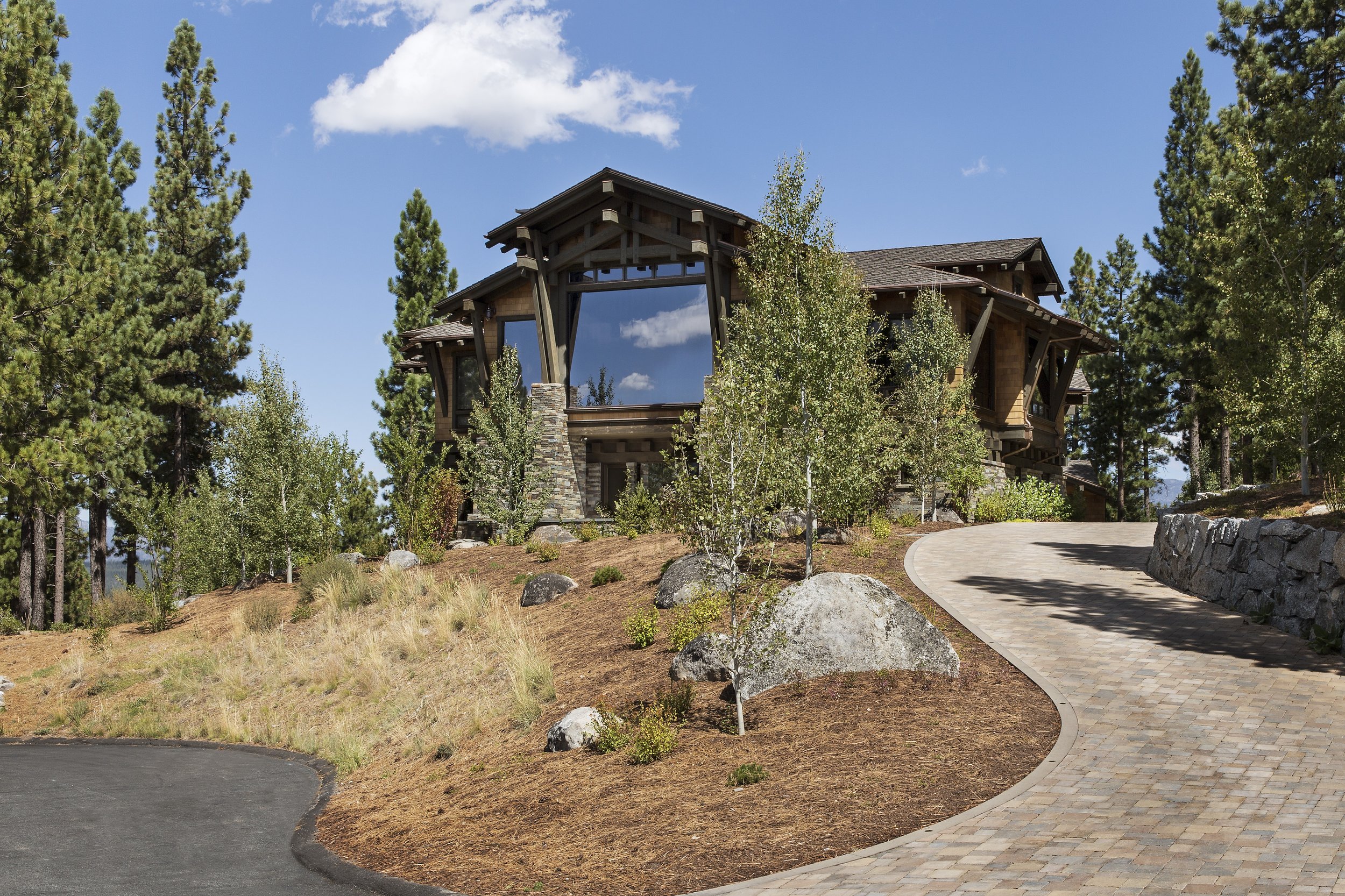 New build project in Tahoe Martis Camp featuring a mountain Craftsman style home inspired by Greene and Greene. The house has a large living room window that brings the outdoors into the space, surrounded by trees and natural landscape.